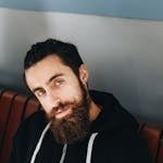 Close-up portrait of a bearded man with green eyes posing indoors against a blue wall.